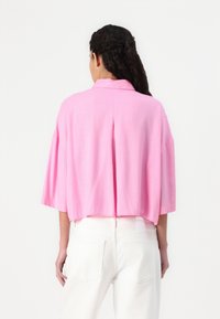 Kortermet, cropped skjorte i lys rosa, med en avslappet passform, teksturert stoff og detalj med ventil bak, kombinert med hvite jeans.