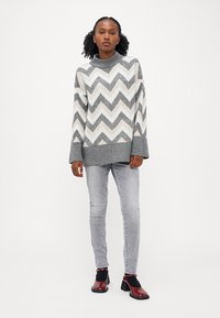 Maglione a righe zigzag grigio e bianco con collo alto, abbinato a jeans grigio chiaro e scarpe con tacco borgogna con accenti neri.