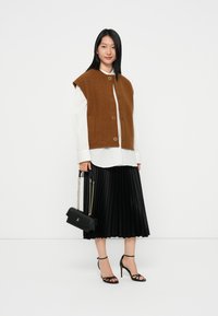 Brauner ärmelloser Pullover über einem weißen Shirt, kombiniert mit einem schwarzen, plissierten Rock. Schwarze Handtasche mit Kettenriemen und schwarzen Riemchenheels.