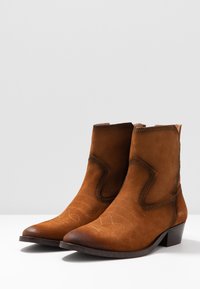Botas de tobillo de ante marrón con punta afilada, patrones de costura sutiles, tacón bajo apilado y paneles laterales elevados. Diseño minimalista.