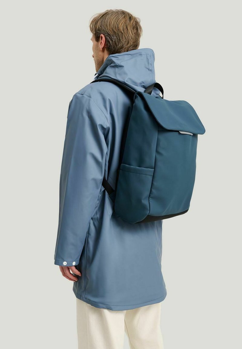 Ryggsäck i mörk teal med en vikbar fäll, tillverkad av ett slitstarkt och smidigt material. Sidoficka synlig. Bärs av en person i en ljusblå kappa.