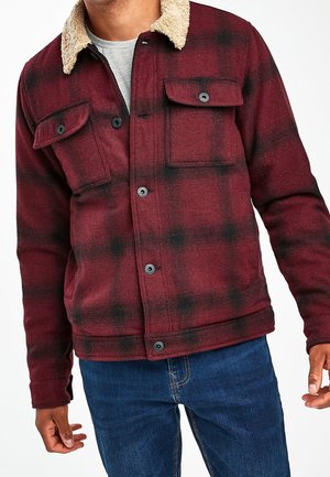 Veste à carreaux rouge et noir avec un col en fausse fourrure beige, fermeture à boutons sur le devant, deux poches poitrine et tissu doux texturé.