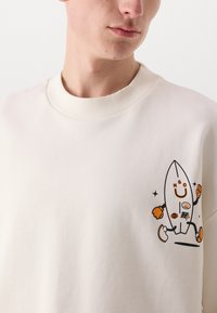 Kremfarget hettegenser med en tegneserie-rocket grafikk med oransje og svarte detaljer, rippekrage og myk tekstur.