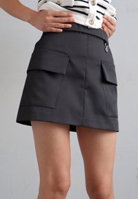 Μαύρη μίνι φούστα με δύο μπροστινές τσέπες, δομημένος σχεδιασμός, λεία υφή και φερμουάρ στη μέση.