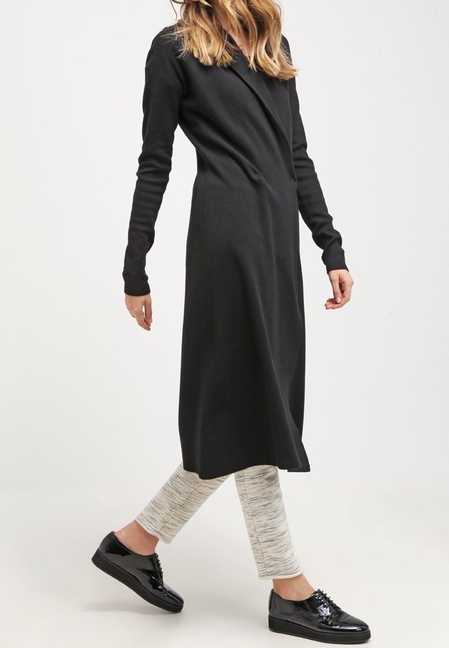 Donna che cammina indossando un vestito midi nero a maniche lunghe sopra pantaloni grigio chiaro a fantasia e scarpe lucide nere con lacci.