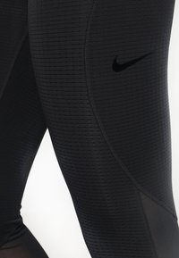 Černé sportovní legíny s texturou a logem Nike na horní části stehna, s síťovinovými panely poblíž lýtek.