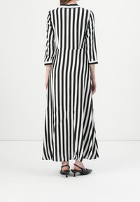 Robe longue à rayures verticales noires et blanches, manches trois-quarts, col en V et ourlet fluide, accompagnée de slingbacks à talons noirs.