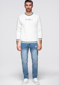 Sweatshirt blanc avec des accents verts et un texte manuscrit, associé à un jean bleu clair déchiré et des baskets blanches. Tenue décontractée.