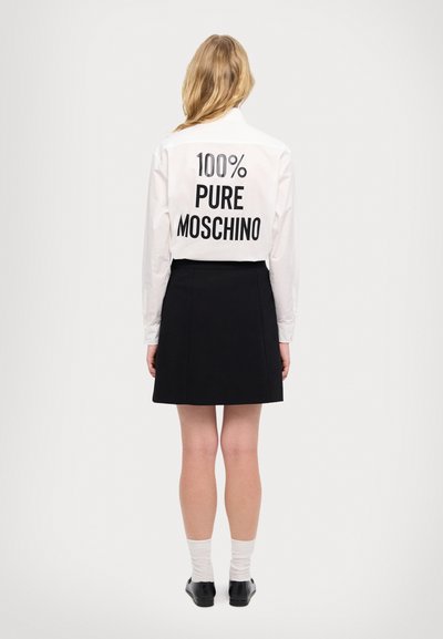 Bela majica z dolgimi rokavi s bold črnim napisom "100% PURE MOSCHINO" na hrbtni strani, v kombinaciji z črno krilom in belimi nogavicami.