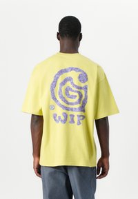 T-shirt en coton jaune avec un grand logo graphique violet au dos, coupe décontractée, manches courtes et col rond, associé à un jean gris.