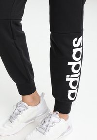 Černé sportovní kalhoty s bílým logem "adidas" na boku, se zúženým střihem a elastickým pasem. V páru s bílými teniskami.