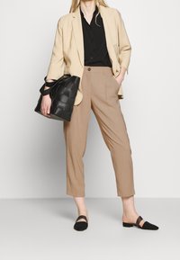 Mujer vestida con pantalones cortos beige, camisa negra, blazer beige, zapatos negros sin cordones, sosteniendo un bolso de mano negro tejido con correa de hombro.