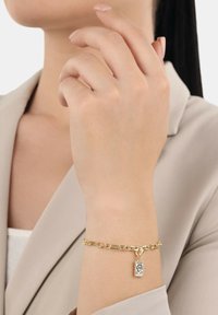 Das Handgelenk einer Frau trägt ein goldenes Kettenarmband mit einem rechteckigen Charme, gekleidet in einem beigen Blazer und einem weißen Oberteil.