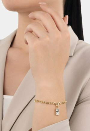 Mujer con una muñeca que lleva una pulsera de cadena dorada con un charm rectangular, vestida con un blazer beige y una blusa blanca.