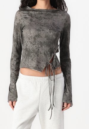 Femme portant un crop top gris à manches longues avec motifs, liens latéraux et pantalon de survêtement gris clair, debout devant un fond uni.