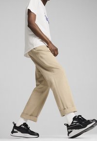 Beige vida byxor med uppvikta fållar, kombinerade med svarta sneakers och vita strumpor, samt en vit t-shirt med kort ärm och blå grafik.