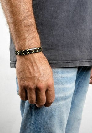 Pulsera trenzada negra y amarilla con un cierre metálico, llevada en una mano con un acabado texturizado, sobre un fondo claro.