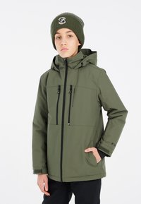 Giacca isolata verde con chiusura zip, due tasche sul petto, cappuccio regolabile e polsini a coste. Abbinata a un berretto lavorato a maglia coordinato.