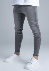 Vékony szabású szürke farmernadrág enyhén kifakult textúrával és látható varrással, fehér sportcipővel párosítva, vastag talppal.