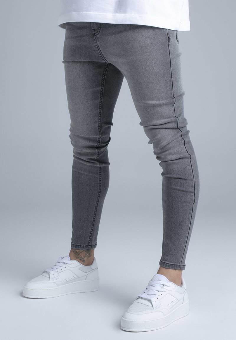 Vékony szabású szürke farmernadrág enyhén kifakult textúrával és látható varrással, fehér sportcipővel párosítva, vastag talppal.