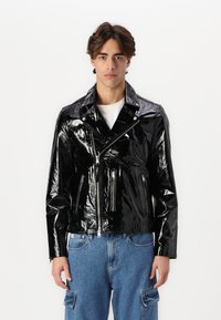 Sort patentlæder motorcykeljakke med cropped design, lynlåslommer og spidse klapper, parret med lyseblå denimbukser.