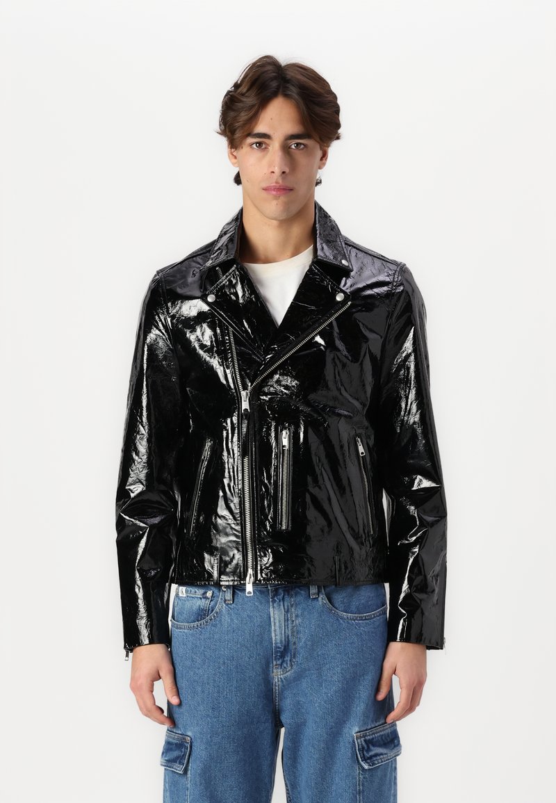 Sort patentlæder motorcykeljakke med cropped design, lynlåslommer og spidse klapper, parret med lyseblå denimbukser.