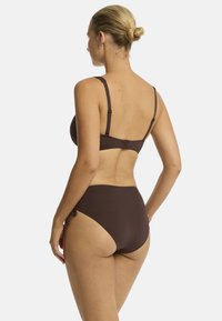Modella che indossa un bikini marrone visto di spalle, con i capelli raccolti in un bun e accessori minimi, su uno sfondo bianco semplice.
