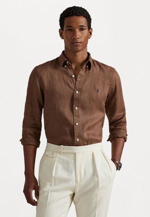 Camicia di lino marrone con maniche arrotolate e polsini abbottonati, con un piccolo logo blu sul petto. Abbinata a pantaloni bianchi a vita alta.