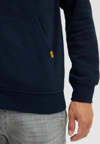 Sudadera de forro polar azul marino con un bolsillo tipo canguro, que presenta una pequeña etiqueta de marca amarilla. Tejido texturizado, puños y dobladillo de canalé.