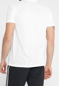 Camisa polo blanca de manga corta con una tela texturizada, con un collar clásico y acentos laterales. Combinada con pantalones negros.