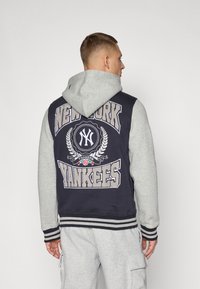 Sudadera con capucha de cuerpo color marino y mangas grises, que presenta un gran logo bordado de los New York Yankees y adornos decorativos en la parte trasera.