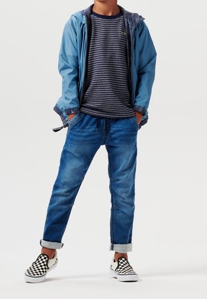 Enfant portant une veste bleue, un t-shirt à rayures bleu marine, un jean retroussé et des chaussures slip-on à carreaux noir et blanc, debout les mains dans les poches.