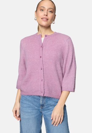 Femme portant un cardigan boutonné violet clair à manches trois-quarts et un jean bleu, debout devant un fond uni.