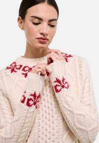 Pull beige clair avec un motif floral rouge sur le yoke et les poignets. Présente un design en tricot torsadé et des ourlets côtelés. Tricot chaud et texturé.