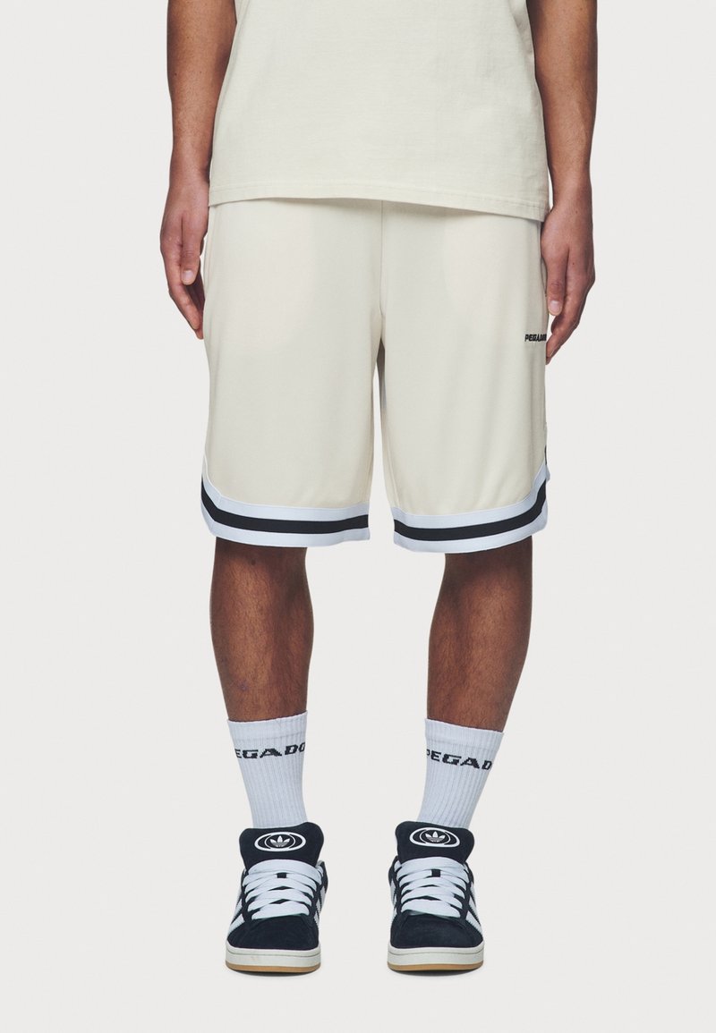 Personne portant un short beige avec un liseré noir et blanc, des chaussettes blanches avec un texte noir, et des baskets noires et blanches, debout.