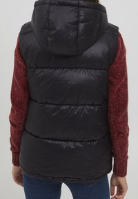 Svart vadderad väst med huva, med en glänsande yta. Ärmarna är i bordeauxfärgad jersey. Designen inkluderar horisontell sömnad och en kroppsnära passform.