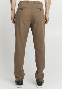 Brown woven trousers with a straight leg design, featuring back pockets with buttons and a clean finish. Accompanied by black sneakers.