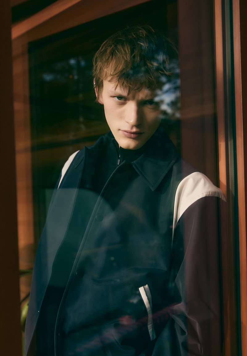 Black and white two-tone jacket with a zip collar, featuring a smooth texture and a front pocket, displayed against a glass window.
