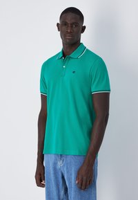 Grünes Polo-Shirt aus Baumwolle, mit einem Kragen in Weiß und einem kleinen Logo. Kombiniert mit hellblauen Denim-Jeans.