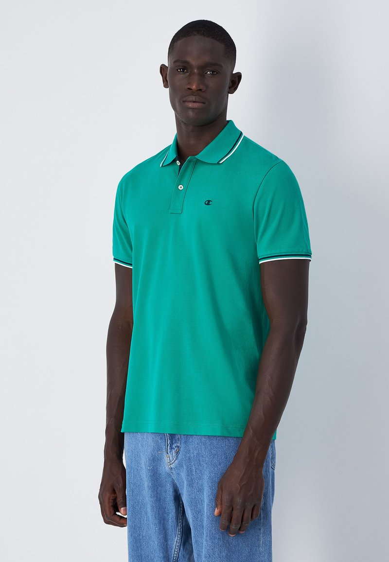 Grünes Polo-Shirt aus Baumwolle, mit einem Kragen in Weiß und einem kleinen Logo. Kombiniert mit hellblauen Denim-Jeans.