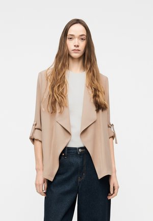 Jeune femme aux longs cheveux ondulés portant une veste beige ouverte avec des manches retroussées, un haut blanc et un jean noir taille haute, sur un fond uni.