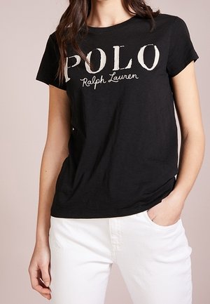 Femme portant un t-shirt noir à manches courtes avec le texte "POLO Ralph Lauren", associé à un pantalon blanc, main dans la poche.