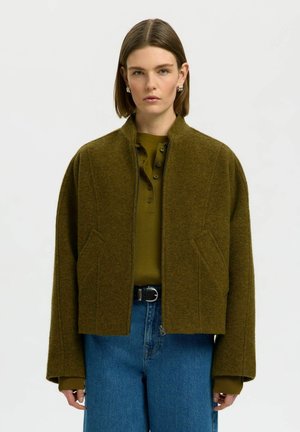 Femme aux cheveux bruns et raides porte une veste en laine vert olive par-dessus un t-shirt henley olive et un jean bleu avec ceinture noire, face à l'avant.