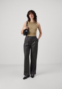 Oliwkowy top na ramiączkach, spodnie o wysokim stanie w czarne paski, płaskie czarne buty. Modelka trzyma okrągłą czarną torbę, stojąc na szarym tle.