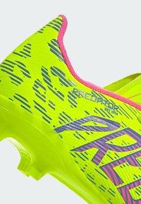 Botas de fútbol de color amarillo brillante con acentos rosas y una parte superior texturizada con líneas azules. Diseño ligero y suela moldeada para una mejor tracción.