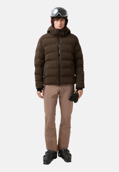 Brown puffer jacket with a hood, zippered front, and side pockets. Paired with light brown ski pants and black ski boots, holding gloves.