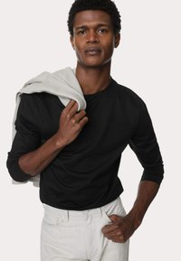 Camisa negra de manga larga, tela ligera, diseño ajustado. Sudadera gris colgada sobre el hombro. Modelo con jeans blancos, pose relajada.