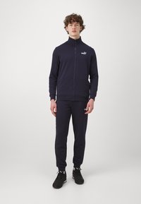Marinblå träningsoverall i mjukt tyg; zip-up jacka med hög krage; matchande byxor med logotyp; bärs med svarta träningsskor.