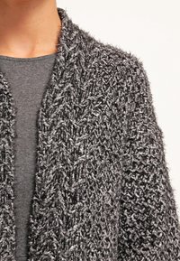 Cardigan grigio a maglia con un motivo testurizzato e un design aperto, indossato sopra un top grigio scuro. Filato morbido e peloso presenta sfumature di colore diverse.
