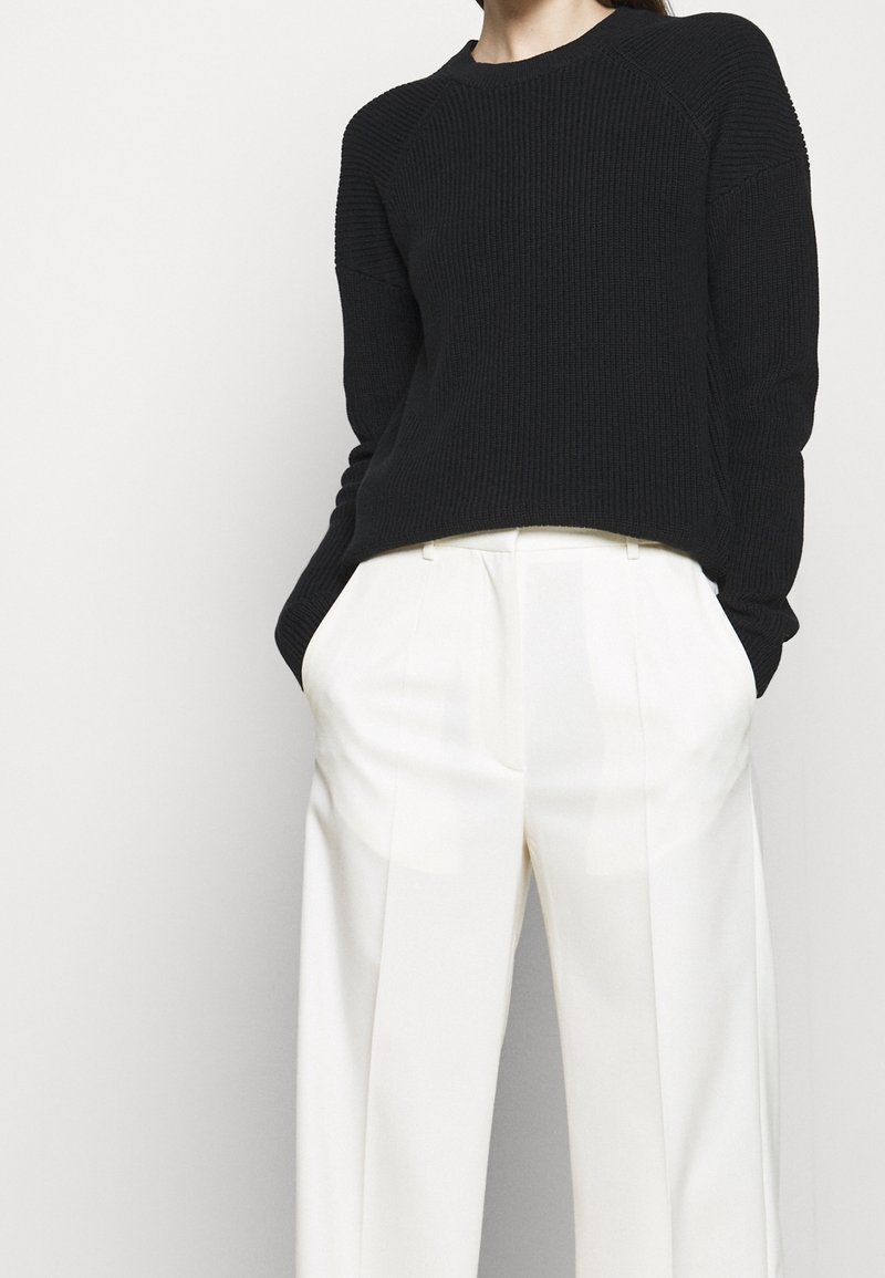 Personne portant un pull côtelé noir rentré dans un pantalon blanc taille haute à jambes larges, les mains dans les poches, sur fond blanc.